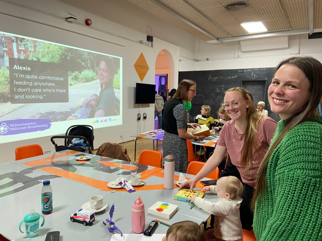 A welcoming community event focused on breastfeeding, featuring women smiling and engaging at a table with children, while a presentation about breastfeeding appears on a screen in the background.